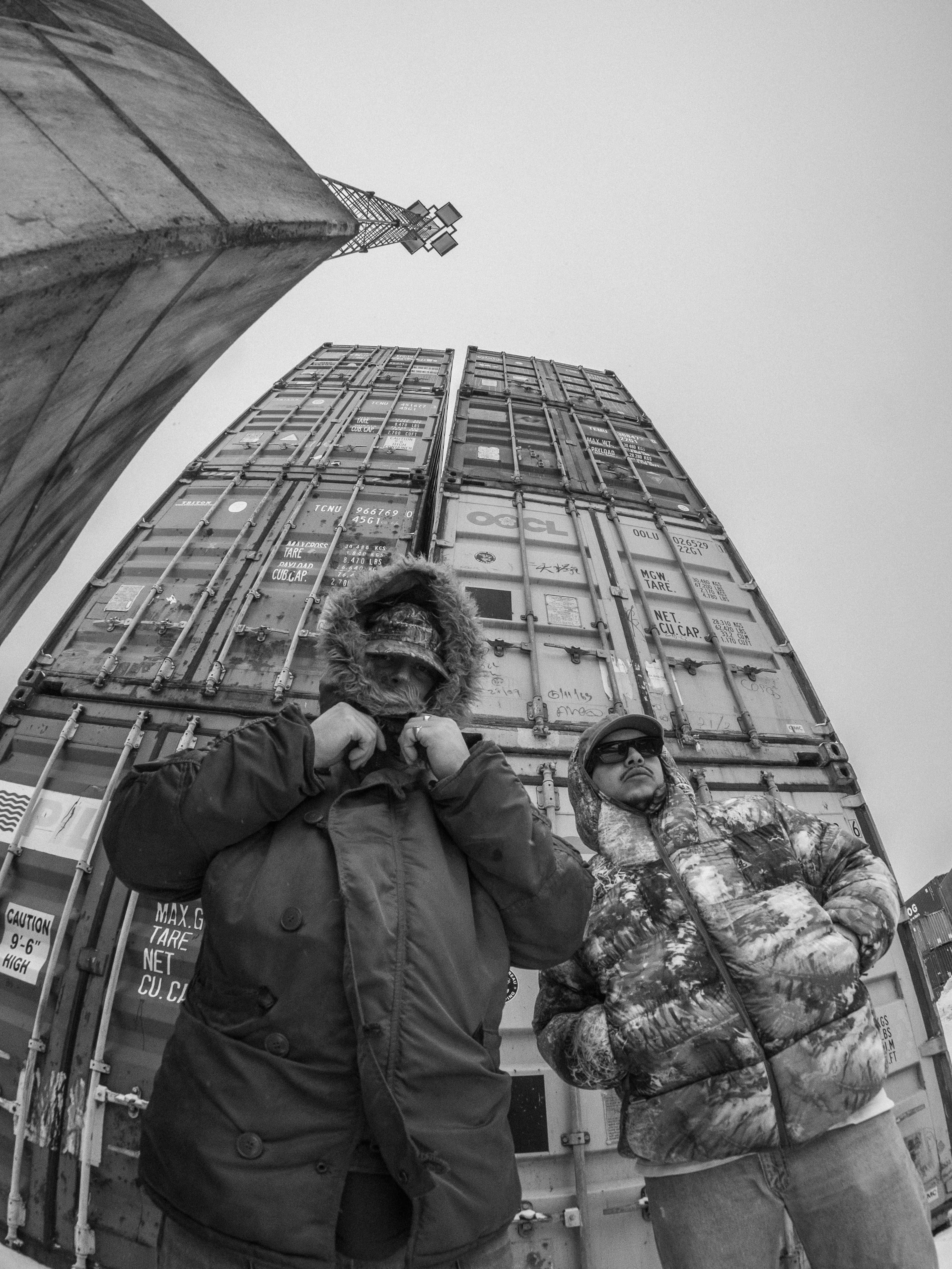 Duoen Tøyen Holding iført varme jakker foran en stabel av shippingcontainere