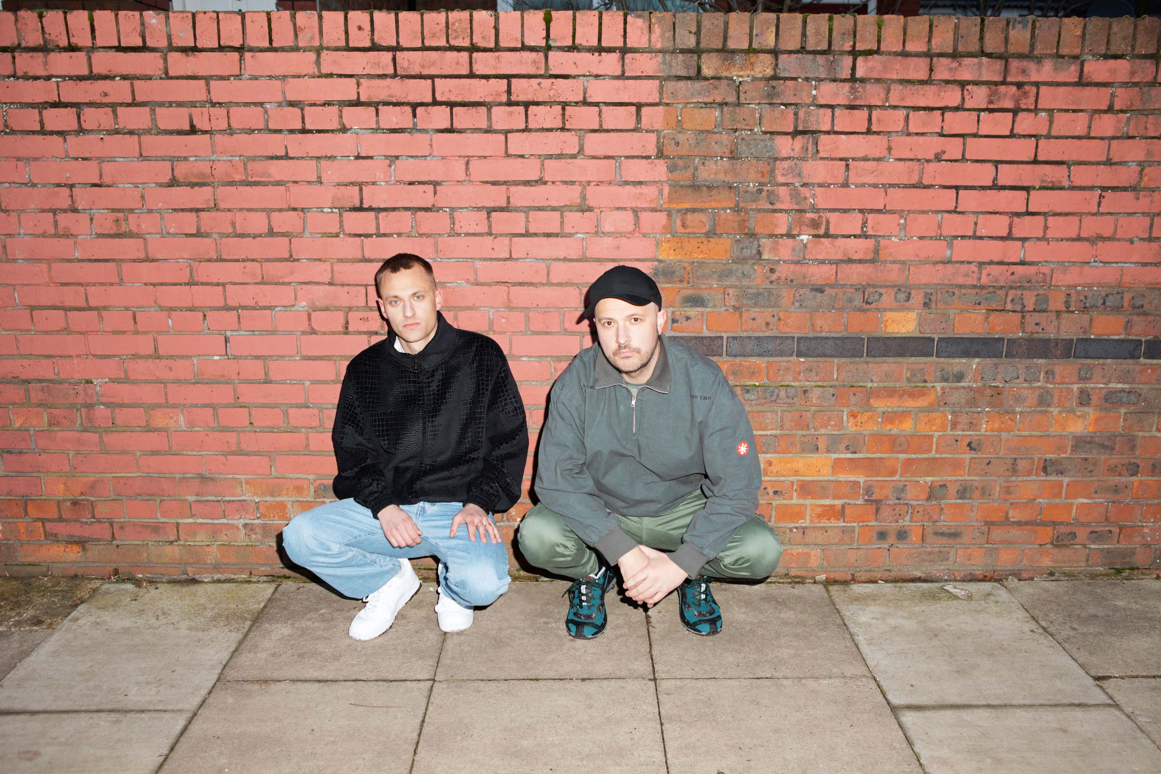 Bilde av den elektroniske duoen Overmono som sitter på huk mot en murvegg